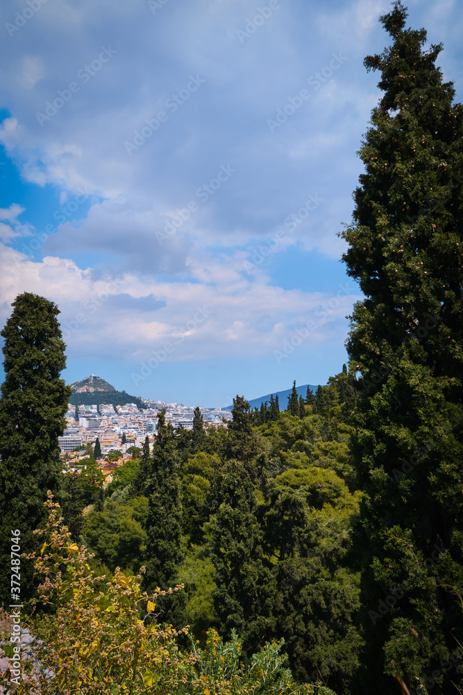 View of Lycabettus hill from Areopagus hill with rich foliage and high cedar or cypress trees. Athens, Greece. Summer day and blue sky with clouds