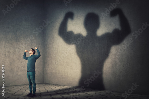 Confident man flexing muscles imagine super power as casting a shadow of muscular bodybuilder showing biceps. Strong person facing his fears. Personal development, inner strength, motivation concept. photo