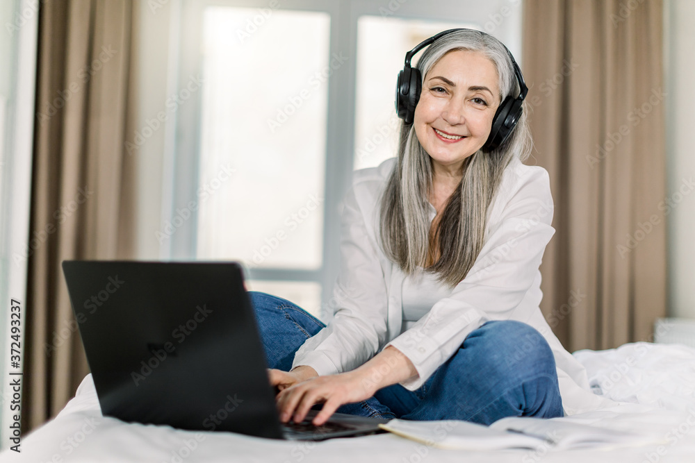 Smiling attractive senior lady with gray hair, sitting on the bed in cozy bedroom at home, listening to music, movie or learning video in headphones and typing on laptop. Smiling attractive senior lady with gray hair, sitting on the bed in cozy bedroom at home, listening to music, movie or learning video in headphones and typing on laptop.