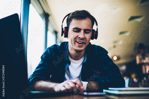 Good looking smiling male student listening to funny music and searching new compositions on smartphone connected to 4G.Young positive man enjoying resting in coffee shop with headphones and device