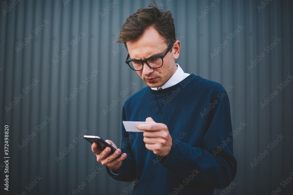 Handsome pensive male blogger dressed in stylish clothes checking visit card and searching information in network via cellular.Young man booking online in website and making payment using service Handsome pensive male blogger dressed in stylish clothes checking visit card and searching information in network via cellular.Young man booking online in website and making payment using service