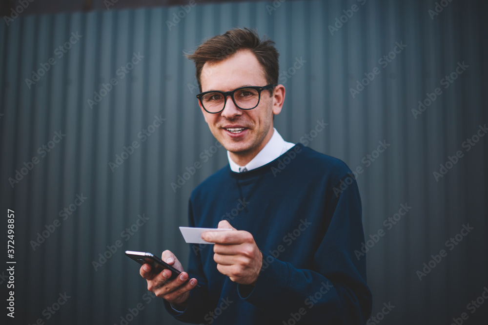 Half length portrait of handsome smiling male student in trendy eyewear enjoying recreation outdoors.Cheerful hipster guy looking at camera while holding visit card and booking online via smartphone Half length portrait of handsome smiling male student in trendy eyewear enjoying recreation outdoors.Cheerful hipster guy looking at camera while holding visit card and booking online via smartphone