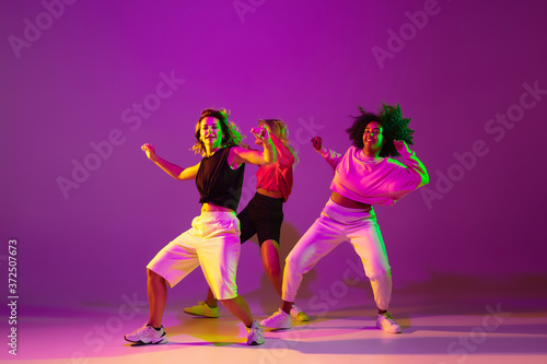 Flexible. Sportive girls dancing hip-hop in stylish clothes on purple-pink background at dance hall in green neon light. Youth culture, movement, style and fashion, action. Fashionable portrait.