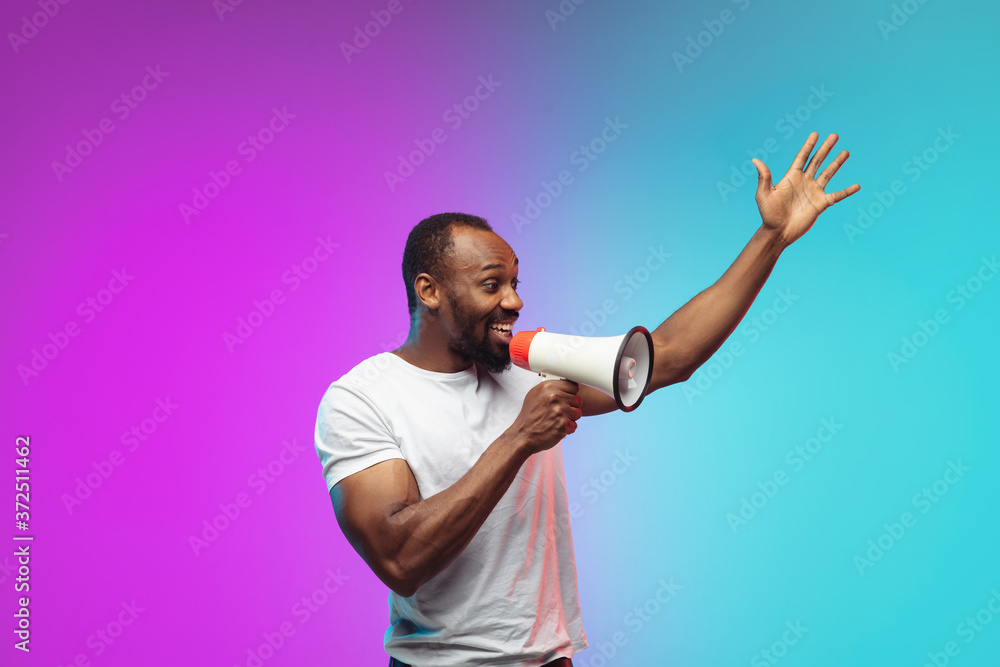Shouting megaphone. African-american young man's portrait on gradient studio background in neon. Beautiful male model in casual style, white shirt. Concept of human emotions, facial expression, sales