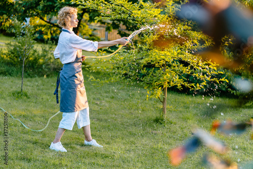 Housewife working in garden. Happy blond woman gardener watering garden with hose. Hobby, leisure concept.