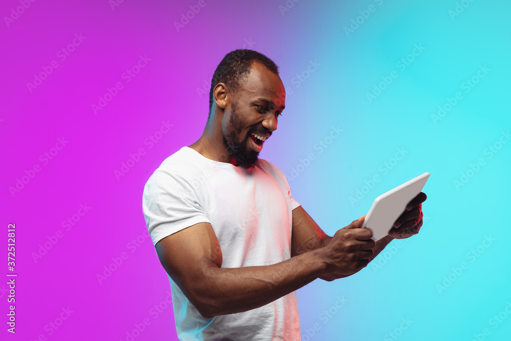 Playing tablet, game. African-american young man's portrait on gradient background in neon. Beautiful male model in casual style, white shirt. Concept of human emotions, facial expression, sales, ad. Playing tablet, game. African-american young man's portrait on gradient background in neon. Beautiful male model in casual style, white shirt. Concept of human emotions, facial expression, sales, ad.