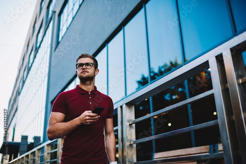 Below view of young man dressed in red t-shirt walking in urban setting with smartphone in hand.Hipster guy in optical eyeglasses strolling in street of modern town and sending sms on telephone