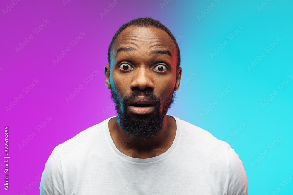 Close up shocked. African-american young man's portrait on gradient background in neon. Beautiful male model in casual style, white shirt. Concept of human emotions, facial expression, sales, ad.