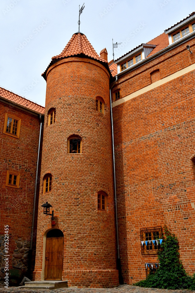 View of a tall round tower of an old medieval castle made out of red brick with many small windows and an angled roof seen during a hike on acloudless summer day on a Polish countryside