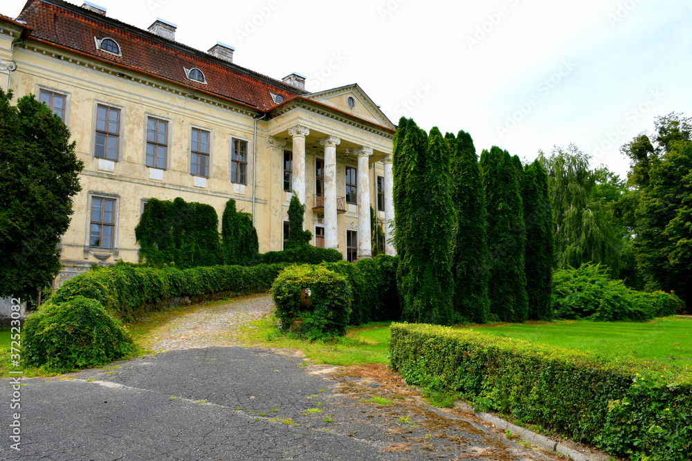 View of an old abandoned and slightly ruined mansion or palace with many windows, arch based entrance, and a slanted red roof surrounded with beautidul gardens, hedges, and tall trees seen in Poland