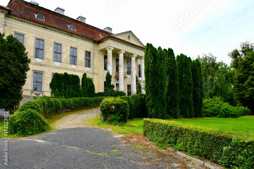 View of an old abandoned and slightly ruined mansion or palace with many windows, arch based entrance, and a slanted red roof surrounded with beautidul gardens, hedges, and tall trees seen in Poland