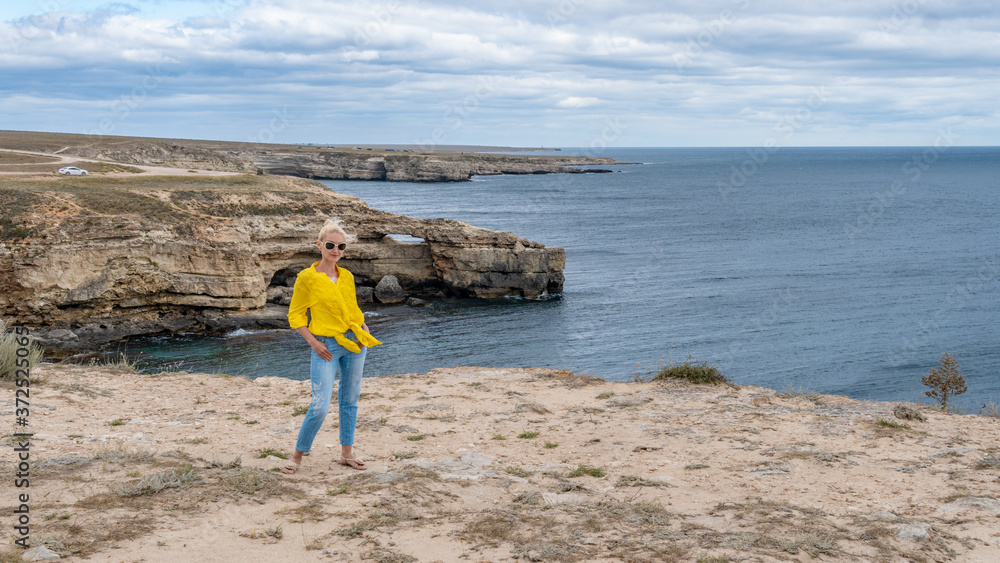 A beautiful girl in a yellow shirt, an adult with a kind look on the background of the sea and rocks, blue clouds during the day and in the summer on a cliff