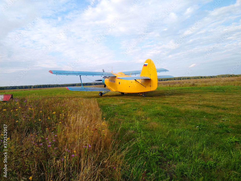 An old yellow biplane with blue wings and a running engine drives through a field of green grass in summer against a blue sky with white clouds An old yellow biplane with blue wings and a running engine drives through a field of green grass in summer against a blue sky with white clouds