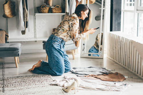 Brunette woman holds smartphone and makes photos of clothes to sell after childbirth via internet. Young woman consults with a designer stylist at home about her wardrobe. photo