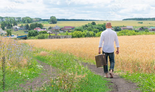 Quadro su tela A brutal young man in a white shirt goes barefoot on the way home holding an old large suitcase