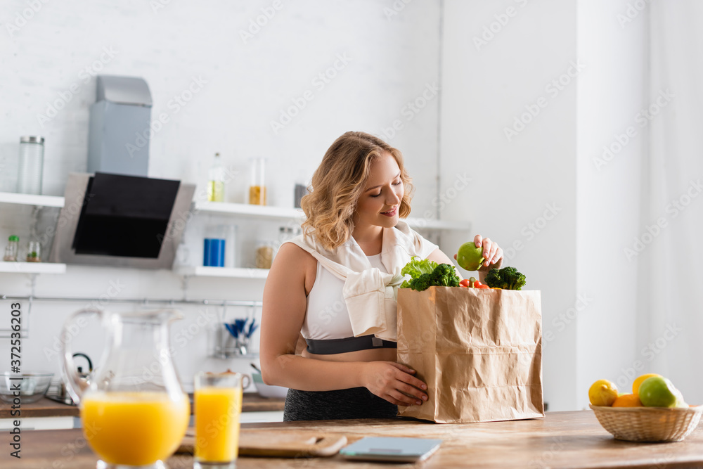 selective focus of young woman holding apple near paper bag with vegetables and fruits in bowl