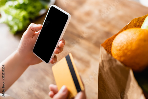selective focus of woman holding smartphone with blank screen and credit card near fruits in paper bag
