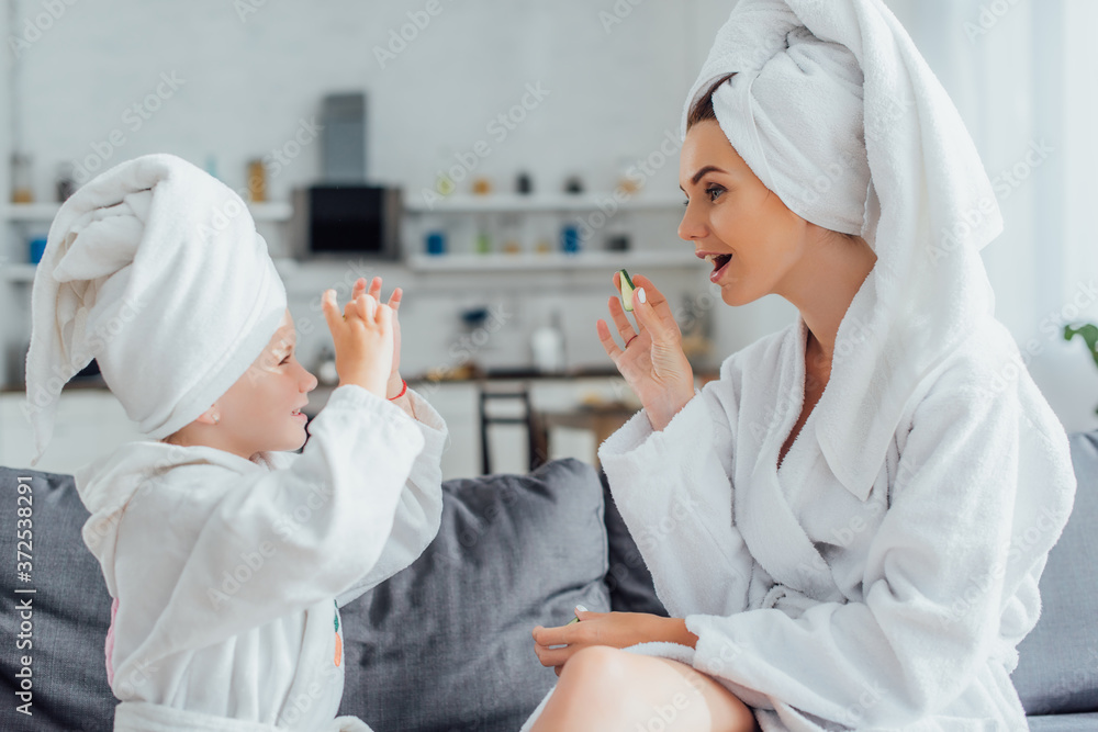 side view of young woman with daughter in white bathrobes and towels on heads holding fresh cucumber slices side view of young woman with daughter in white bathrobes and towels on heads holding fresh cucumber slices