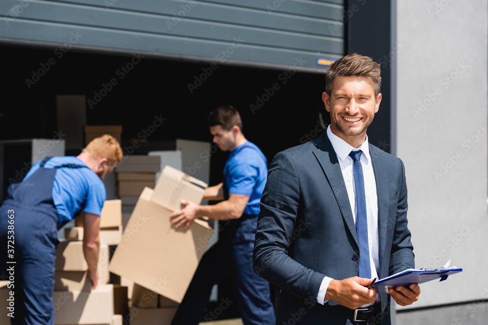 Selective focus of businessman with clipboard looking at camera while movers working in warehouse outdoors Selective focus of businessman with clipboard looking at camera while movers working in warehouse outdoors