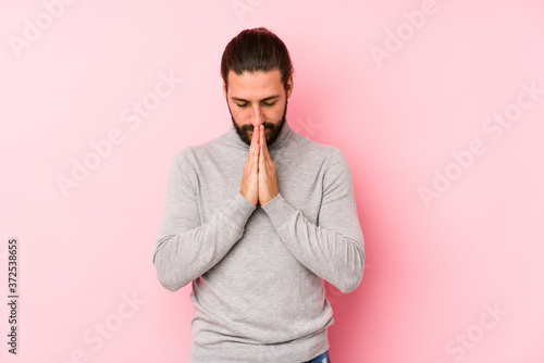 Young long hair man isolated on a pink background holding hands in pray near mouth  feels confident.