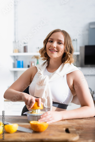 selective focus of young woman squeezing orange in juicer while preparing juice in kitchen