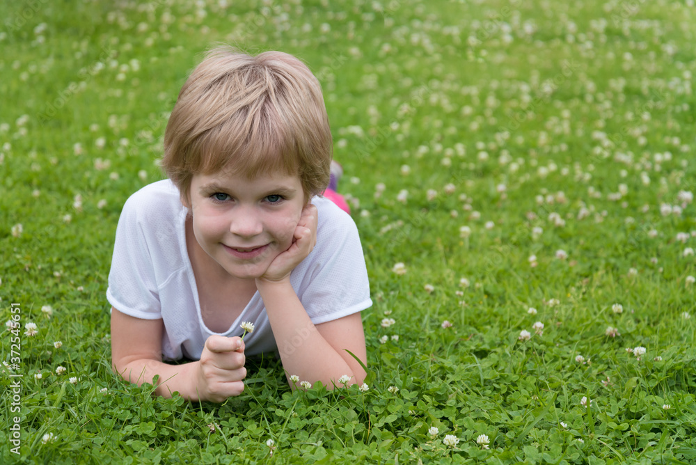 The child is lying on a green lawn. The blond boy smiles and looks at the camera. A blonde girl surrounded by clover flowers. Space for text. The child is lying on a green lawn. The blond boy smiles and looks at the camera. A blonde girl surrounded by clover flowers. Space for text.