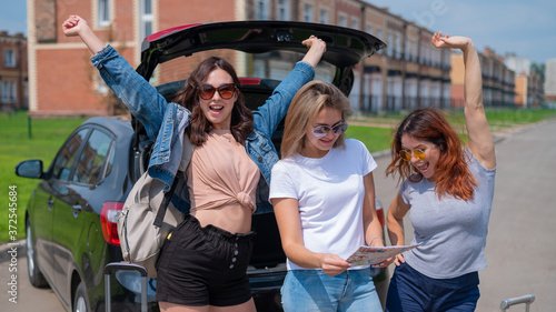 Three friends are going on a road trip for the summer vacation and are looking at the map. Young women load suitcases into the trunk of a car.