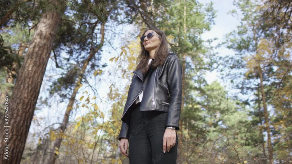 4K portrait of confident woman in black leather jacket and cool sunglasses standing in the middle of gorgeous autumn forest. Low angle 360 degree tracking arc shot. 4K portrait of confident woman in black leather jacket and cool sunglasses standing in the middle of gorgeous autumn forest. Low angle 360 degree tracking arc shot.