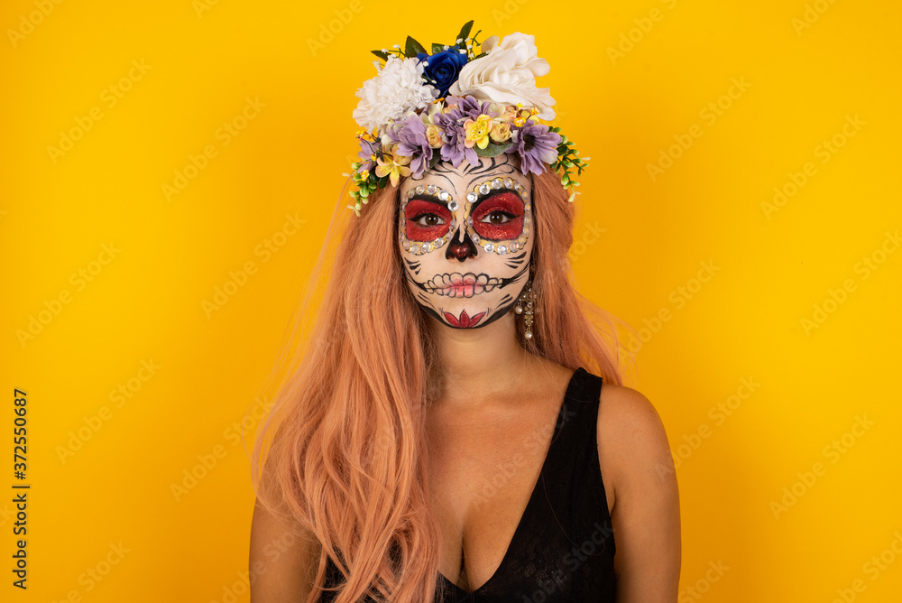 Waist up shot of joyful young woman wearing halloween make up looking to the camera, thinking or wondering about something. Both arms down, neutral facial expression.