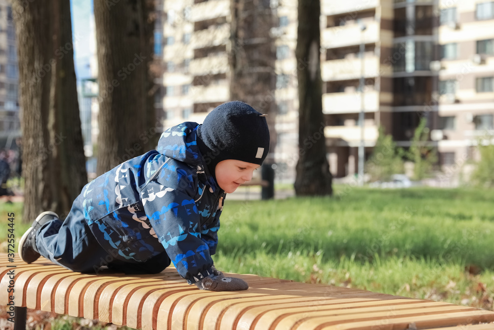Little boy funny crawls on a wooden bench in the park. He's wearing a jumpsuit, hat and mittens. The boy has fun. Little boy funny crawls on a wooden bench in the park. He's wearing a jumpsuit, hat and mittens. The boy has fun.