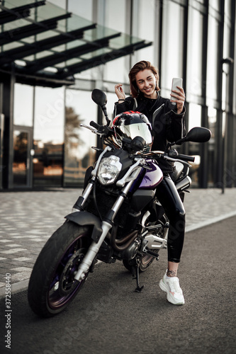 Smiling girl in a motorcycle jacket sits on a purple motorbike and looks at her phone