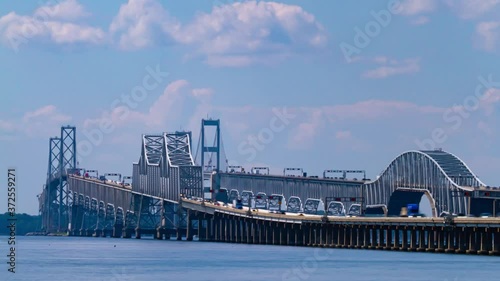 A day time telephoto time lapse footage showing the rush hour traffic on Chesapeake Bay Bridge. It features detailed view of the bridge with columns and suspensions as well as the boats passing under.