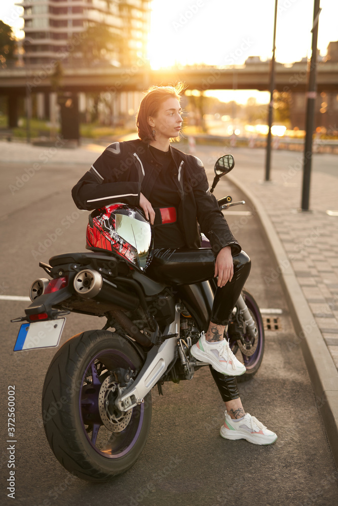 Very beautiful girl in a black motorcycle jacket sits with red and black motorcycle helmet on a black sport motorbike