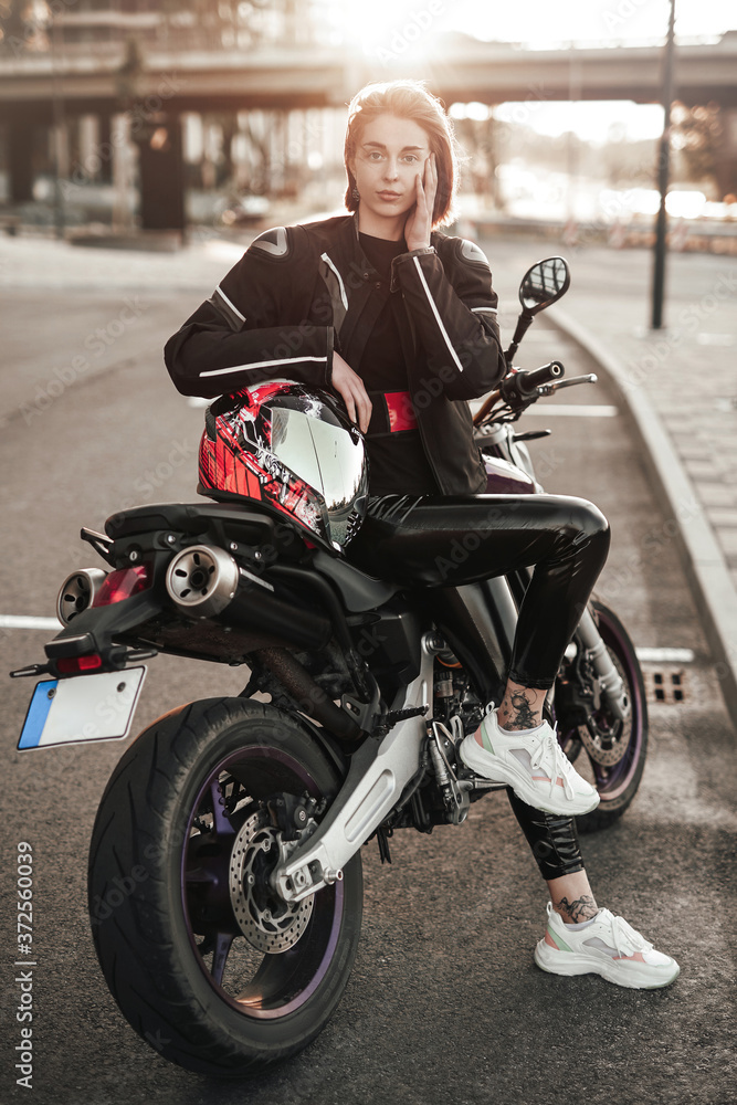 Beautiful young brunette girl in a black motorcycle jacket sits on a purple sport motorbike, with the sunset on the background