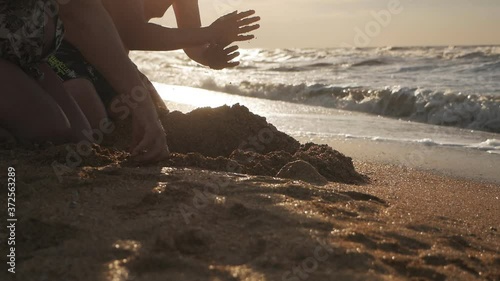 On a summer evening, two men kneel down and dig a hole with their hands on the beach by the sea. Men are funny as children play on a sandy beach in a desert area by the ocean