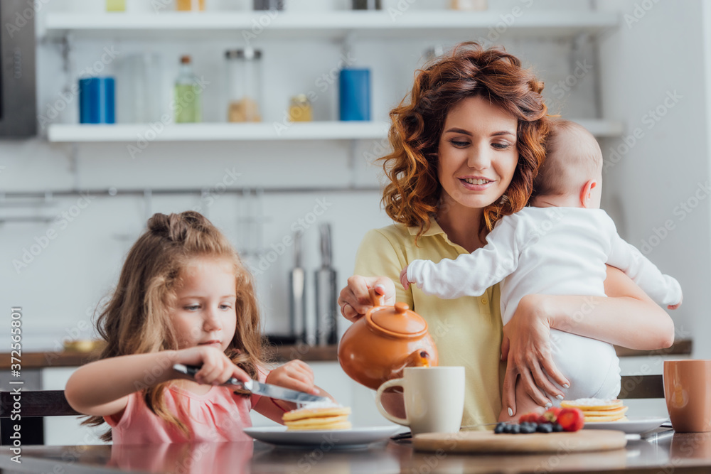 selective focus of mother pouring tea from teapot while holding infant near daughter eating pancakes selective focus of mother pouring tea from teapot while holding infant near daughter eating pancakes