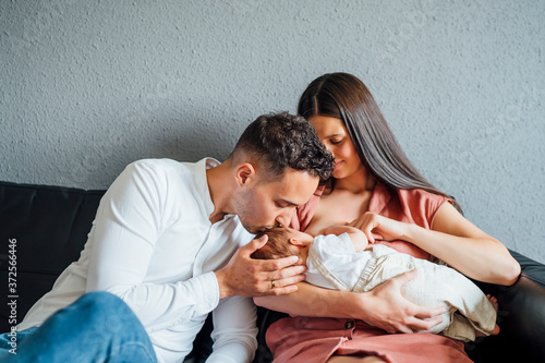 Happy father kissing baby boy being breastfed by mother at home