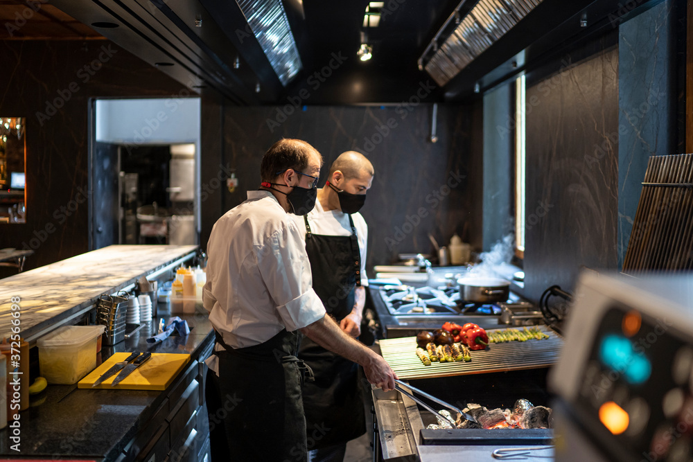 Chefs wearing protective face masks preparing grilled vegetables in restaurant kitchen Chefs wearing protective face masks preparing grilled vegetables in restaurant kitchen