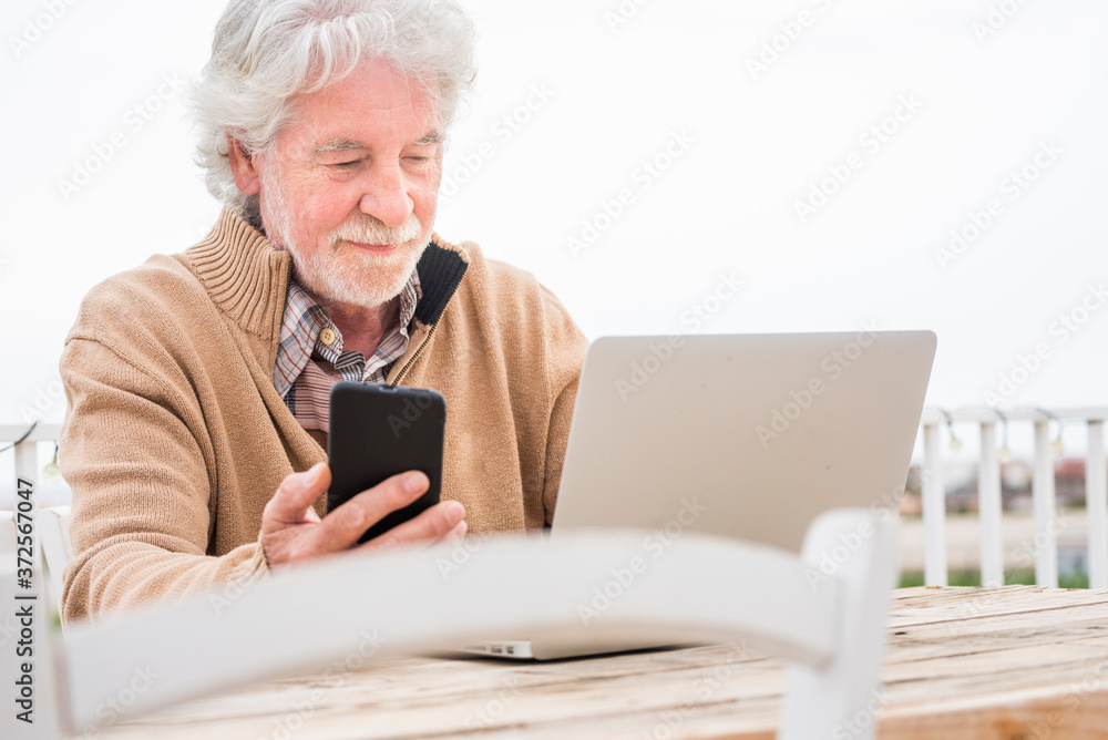Senior man holding mobile phone while using laptop at table Senior man holding mobile phone while using laptop at table