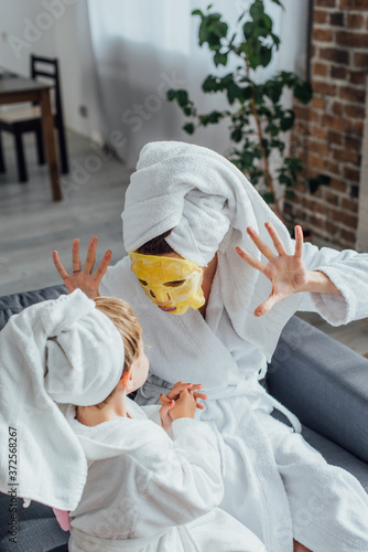 Quadro su tela high angle view of mother in face mask showing scaring gesture while sitting wit