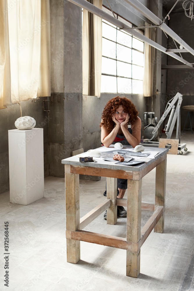 Female stonemason with hands on chin leaning by sketches and work tools over workbench in studio Female stonemason with hands on chin leaning by sketches and work tools over workbench in studio