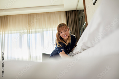 Smiling chambermaid making bed while standing in hotel room