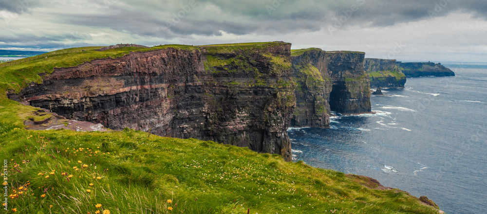 Cliff of Moher, county Clare, Ireland. Famous Irish tourist destination. Panorama image. Grey storm sky. Blue ocean and green grass with flowers. Cliff of Moher, county Clare, Ireland. Famous Irish tourist destination. Panorama image. Grey storm sky. Blue ocean and green grass with flowers.