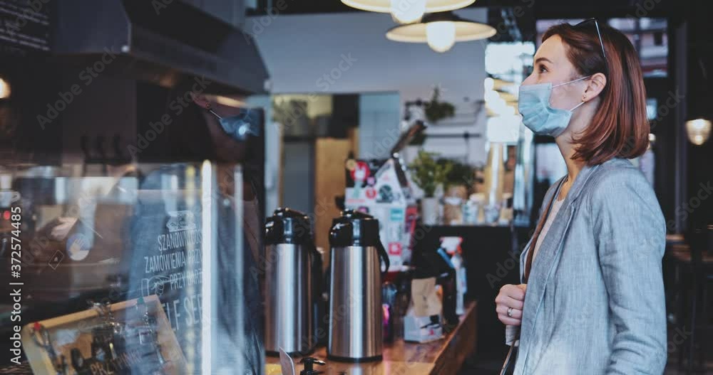 Woman Customer In Protective Mask Ordering In A Coffeeshop. SLOW MOTION, Stabilizer. BusinessWoman in face mask against air pollution and coronavirus paying for coffee with contactless credit card. Woman Customer In Protective Mask Ordering In A Coffeeshop. SLOW MOTION, Stabilizer. BusinessWoman in face mask against air pollution and coronavirus paying for coffee with contactless credit card.