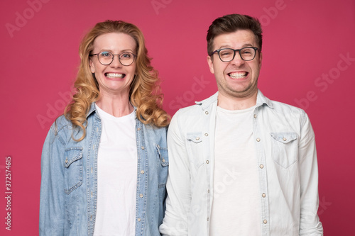 Funny caucasian woman and man smiling happily while looking at camera, standing over pink background