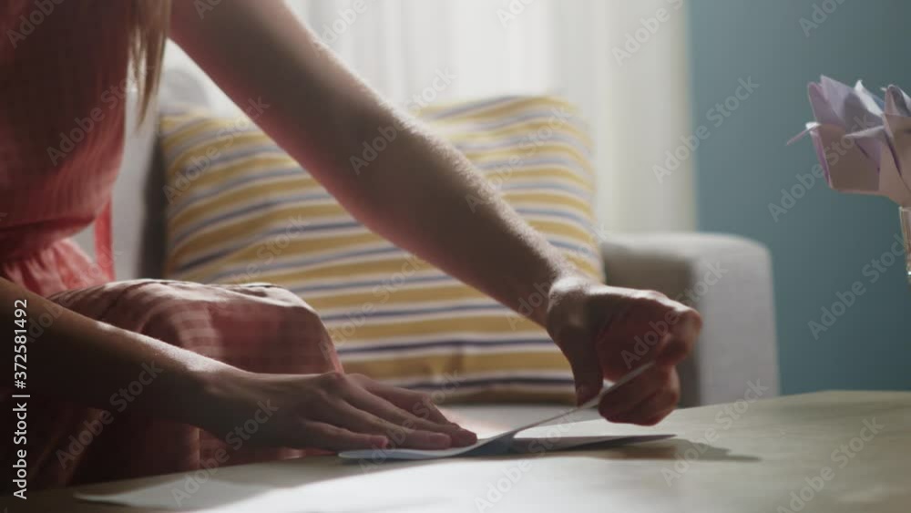 Young girl girl in a pink dress sits on the couch and folds origami from a white sheet of paper on a coffee table. Close-up Young girl girl in a pink dress sits on the couch and folds origami from a white sheet of paper on a coffee table. Close-up