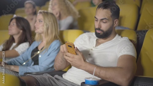 Handsome young Middle Eastern man using smartphone in cinema with annoying Caucasian women talking at the background. Portrait of irritated film-lover distracted from film in movie theatre. photo