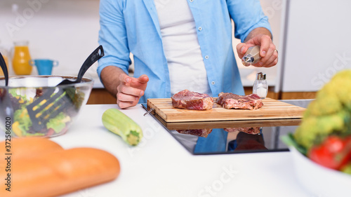 Chef pours salt on pork or beef meat with salt shaker at home cozy kitchen. Barbecue concept and tasty food lifestyle.