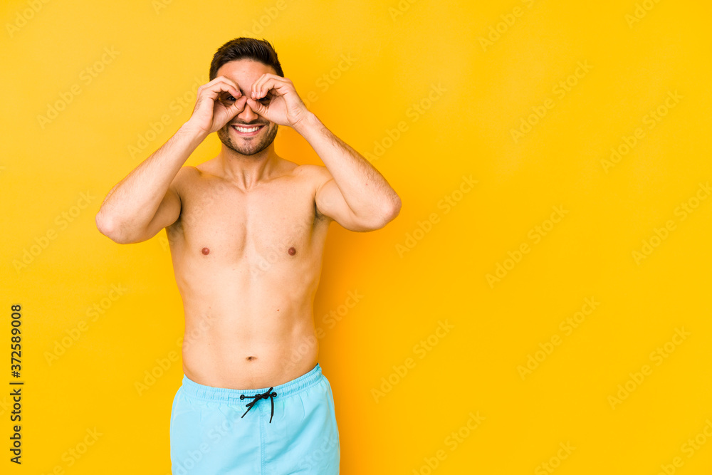 Young caucasian man with swimsuit isolated on yellow background Young caucasian man with trshowing okay sign over eyes