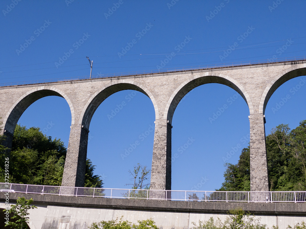 gigantic impressive railway viaduct without train in front of blue sky and green forest with a highway and guardrails by day without people